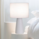 Ingá Table Lamp by Geo Contemporary in White with a White shade, lit, beside a bed