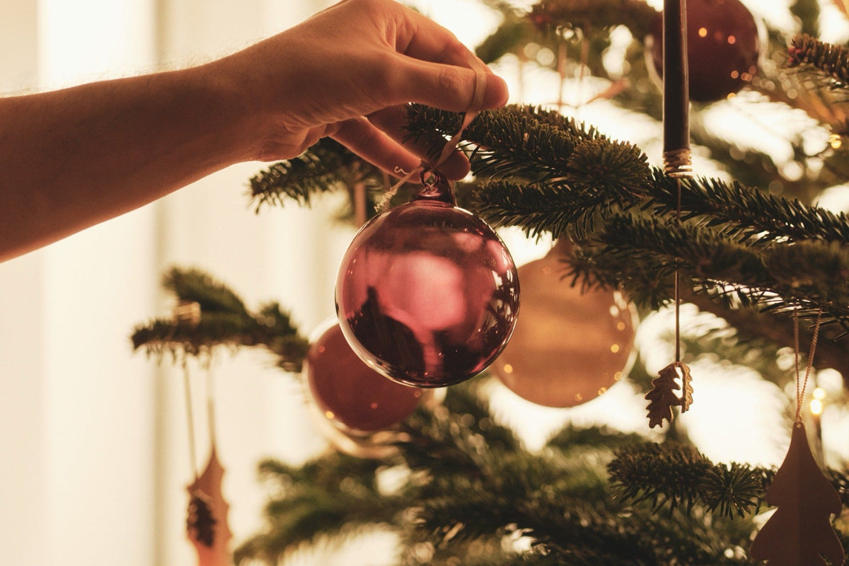 Hand hanging a deep red glass bauble on a Christmas tree decorated with brass oak leaf ornaments in warm afternoon light