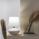 Cedro Table Lamp by Geo Contemporary with a White shade, lit, on a console beside dried grasses