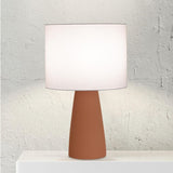 Ingá Table Lamp by Geo Contemporary in Terracotta with a White shade, lit, against a white brick wall